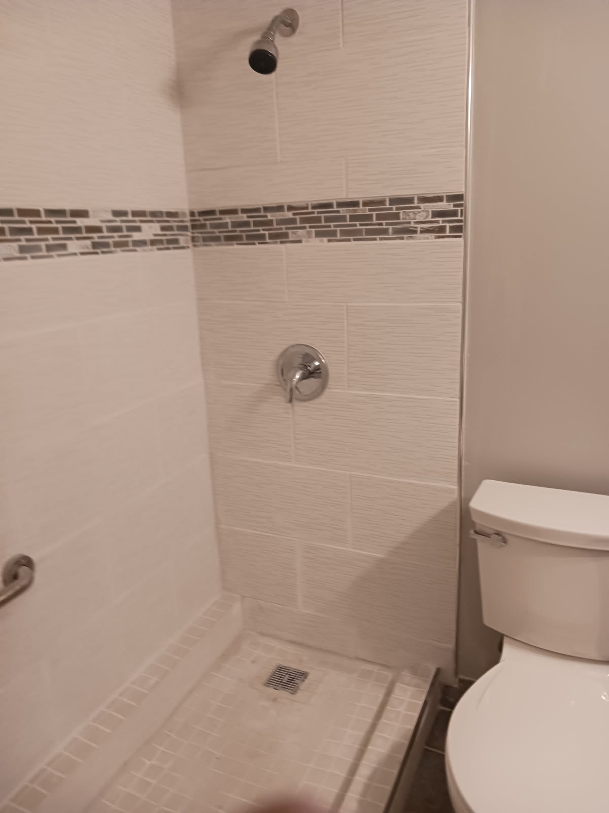 Walk-in shower finished in white textured wall tile with a decorative mosaic strip, a chrome shower head and a square floor drain