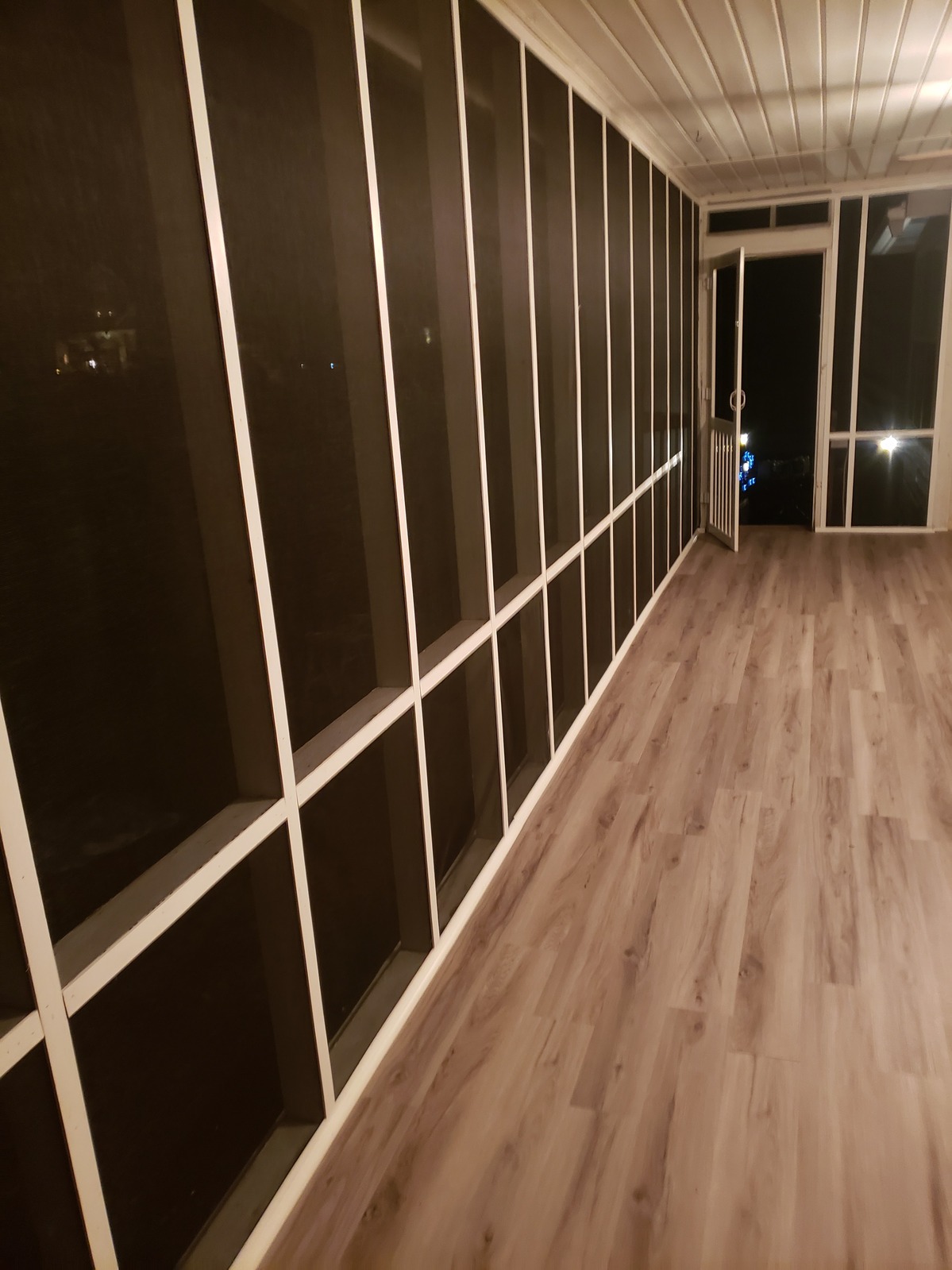 Enclosed porch corridor with a white-framed grid of dark glazed panels on one side and a glass door opening to the night