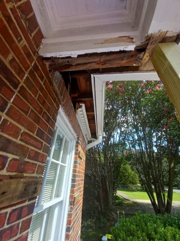 Corner of a brick house showing rot and damage to the wooden roof eaves and soffit, with the roof framing exposed above a window