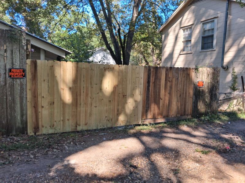 Wooden fence line with a weathered gate section, a newer light plank section, and older dark boards, on a gravel yard under trees