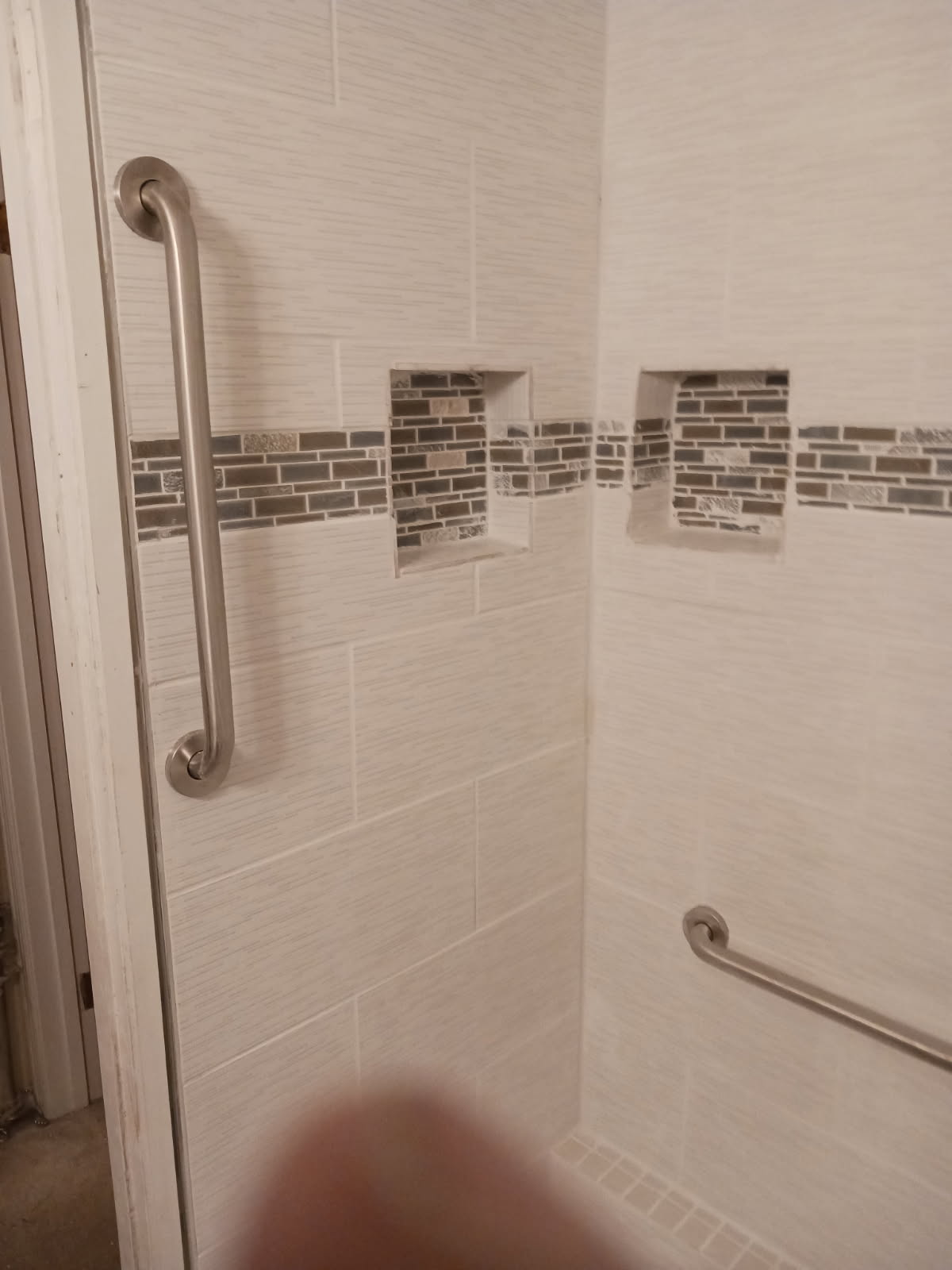 Inside corner of a tiled shower with a mosaic accent band running through two recessed niches and two brushed metal grab bars