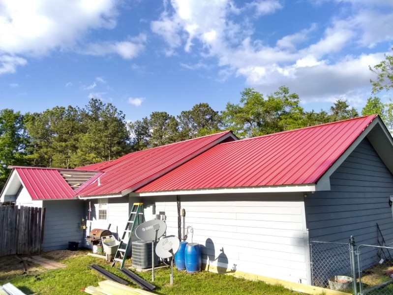 Finished red metal roof on a single-story building with light gray siding, against a line of green trees