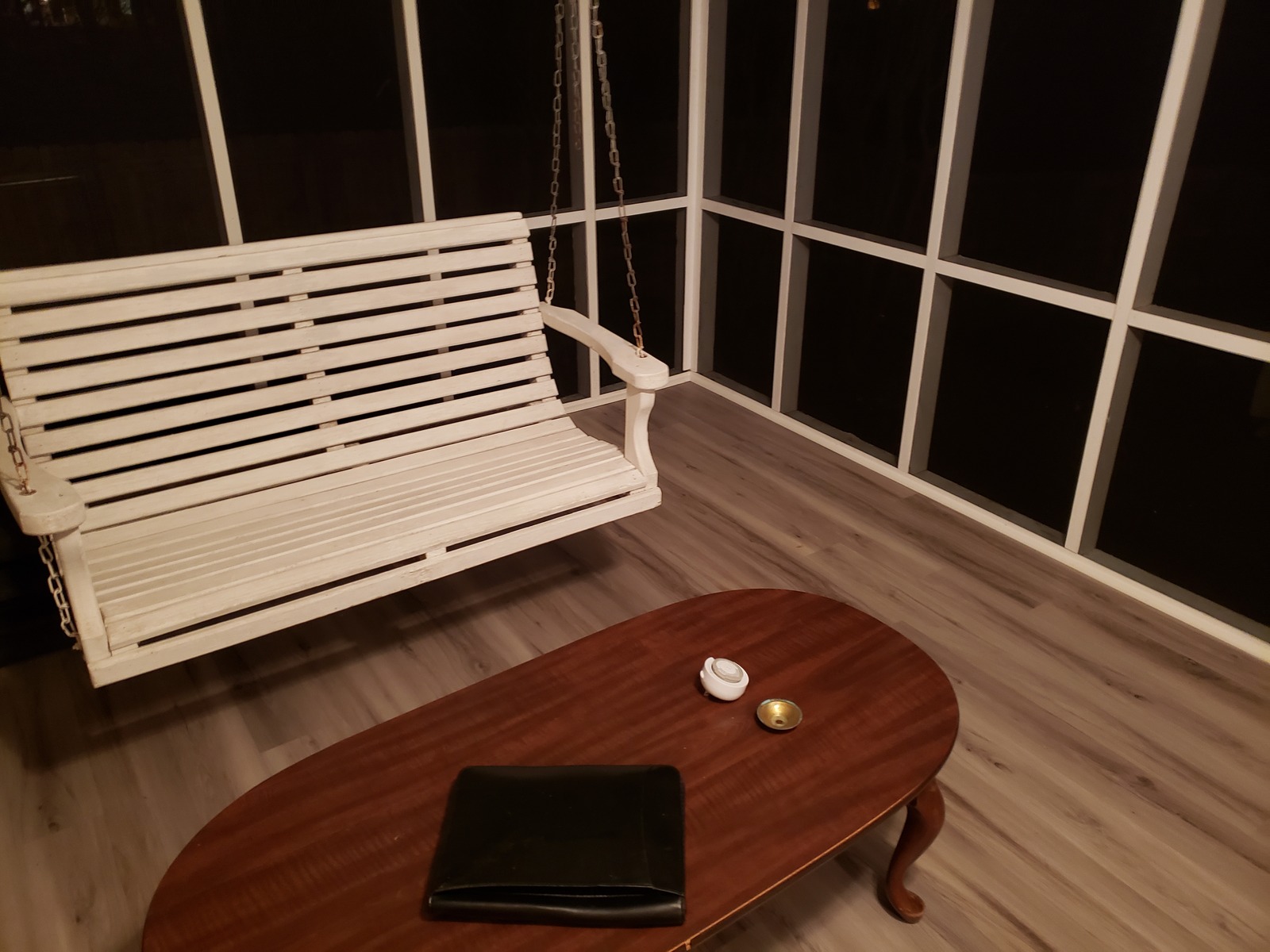 White slatted wooden porch swing hung by chains over light plank flooring, with an oval coffee table in front of it