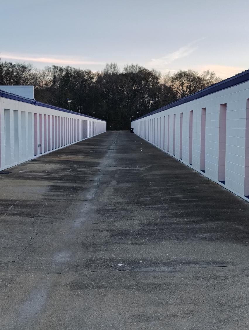 Long concrete driveway running between two rows of single-story storage units with pale brick walls and colored door panels