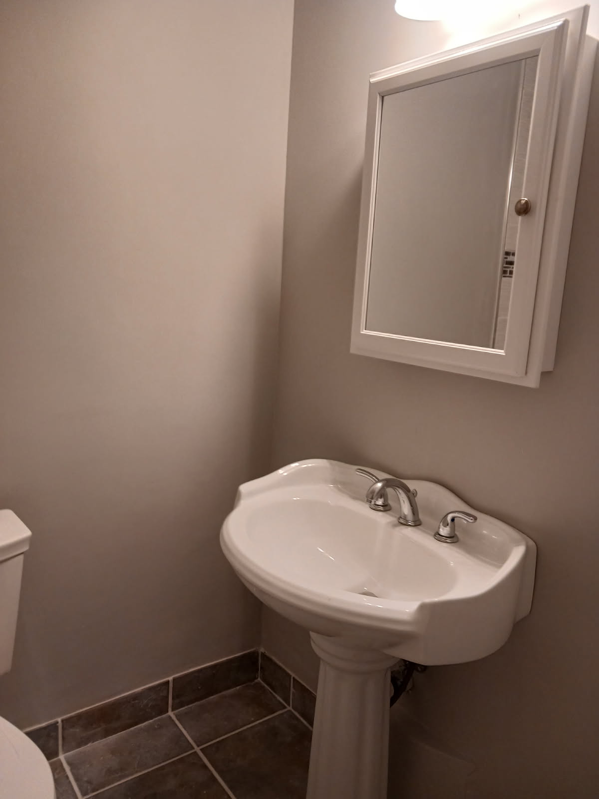 Bathroom corner with a white pedestal sink and chrome faucet below a mirrored cabinet, on a dark tiled floor