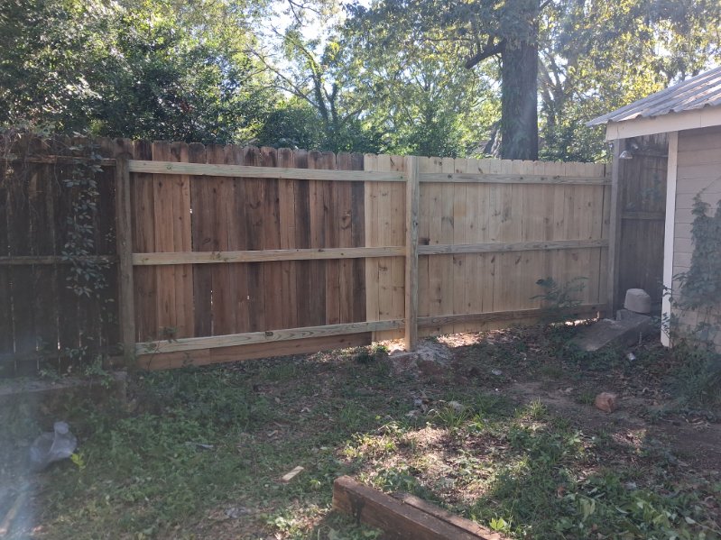 Wooden privacy fence in a yard with an older dark section beside two newer light plank sections and trees behind
