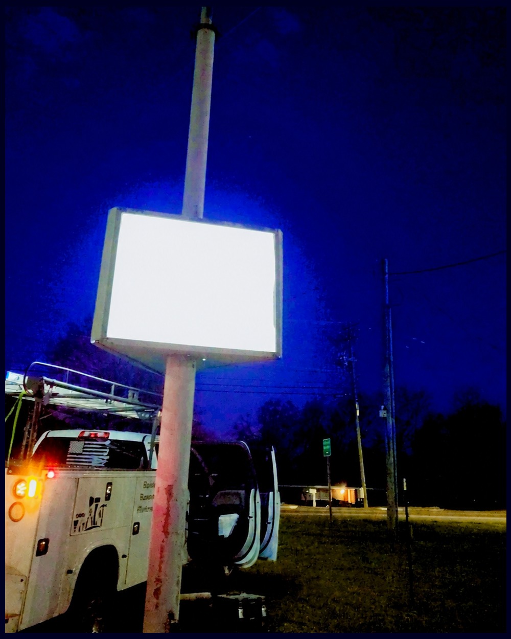 White work truck with a ladder rack parked at night in front of a lit commercial storefront sign