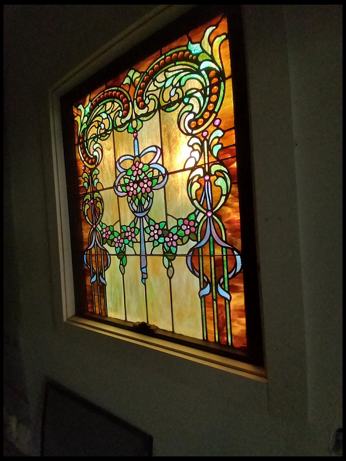 Rectangular stained glass window with a floral and vine design in pink, green and blue glass, lit from behind and set into a dark wall