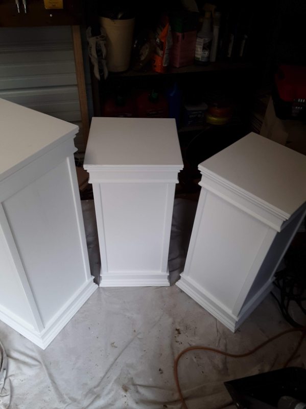 Three white painted wooden cabinet pieces with paneled sides standing on a drop cloth in a workshop