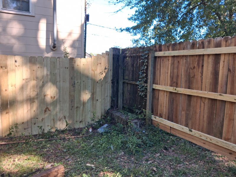 Backyard fence where a newer light plank section abuts an older weathered section, with a house and downspout behind